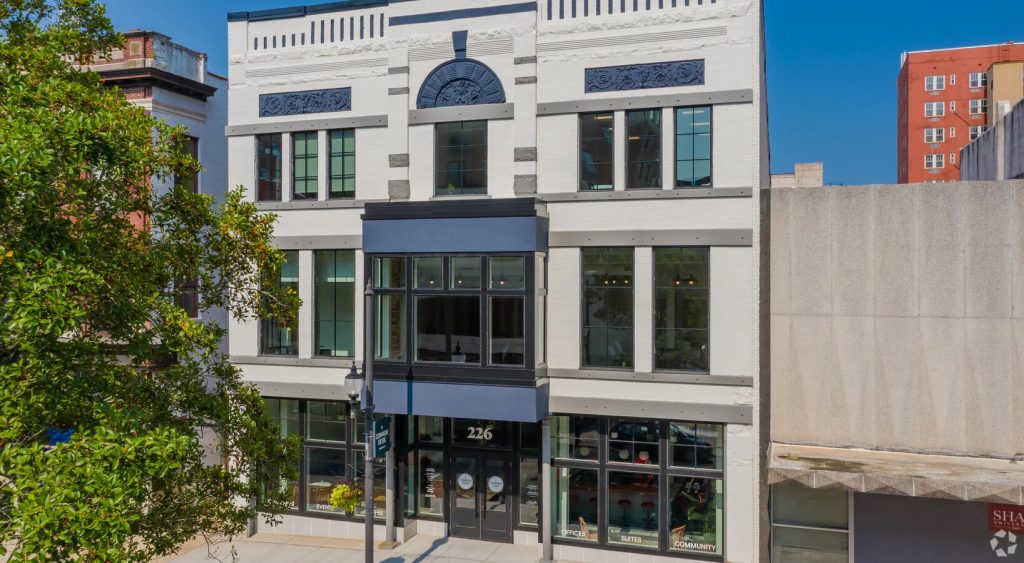 A historic three-story building with a white facade, dark accents, and large windows. A tree partially frames the facade. The atmosphere is vibrant and urban.